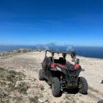 Red and black off-road vehicle parked on rocky mountaintop overlooking blue ocean and coastal landscape