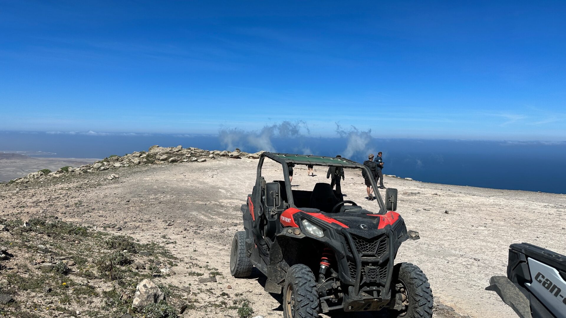 Red and black off-road vehicle parked on rocky mountaintop overlooking blue ocean and coastal landscape