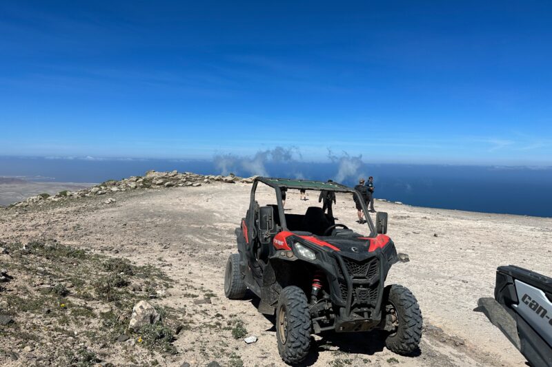 Red and black off-road vehicle parked on rocky mountaintop overlooking blue ocean and coastal landscape