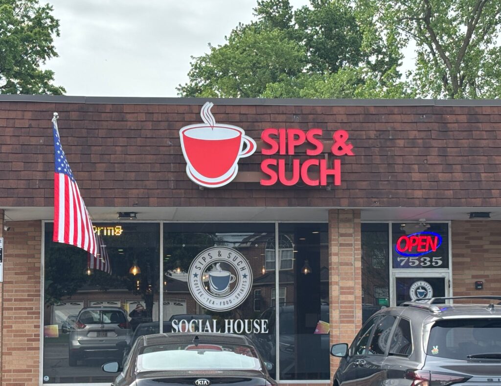 Brick storefront for "Sips & Such" coffee shop displaying an American flag and open sign with parked vehicles outside