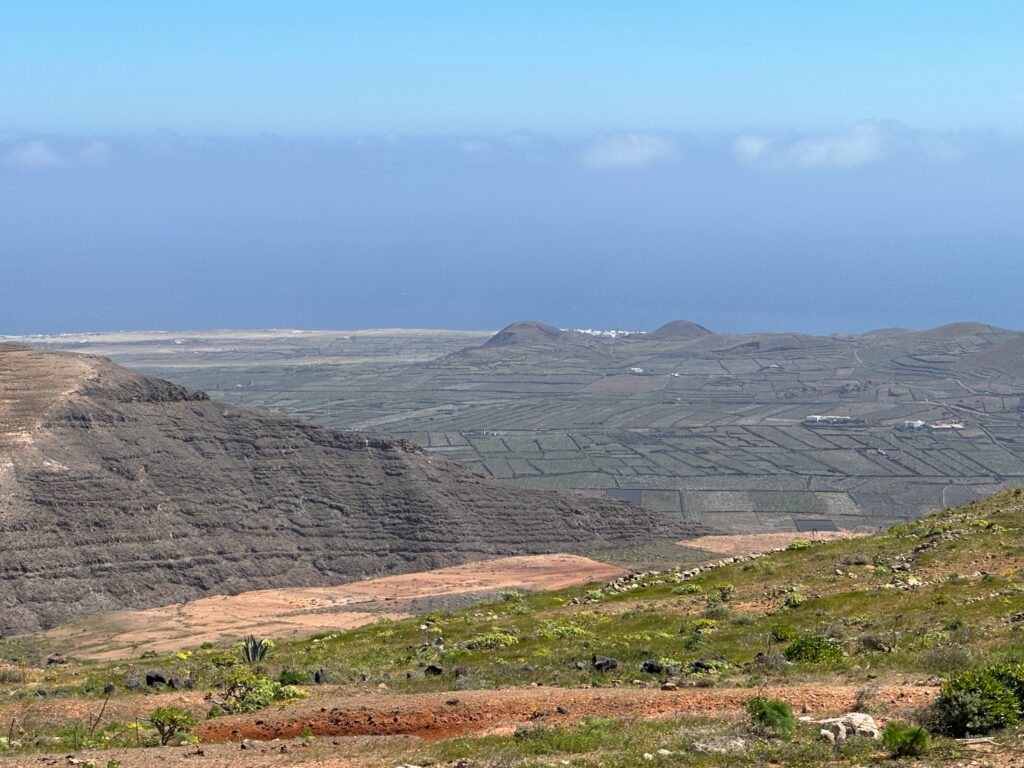 Volcanic landscape with terraced agricultural fields, red soil, and distant mountains under blue sky