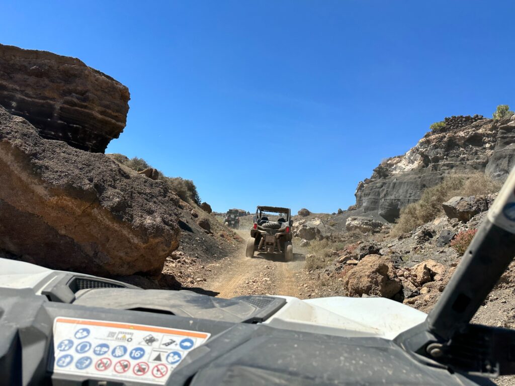 Two off-road vehicles navigate a rocky desert trail surrounded by large boulders under clear blue sky