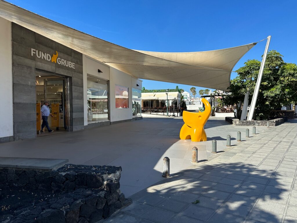 Modern plaza with FUNDA GRUBE storefront, yellow sculptural art installation, and white fabric canopy under clear blue sky