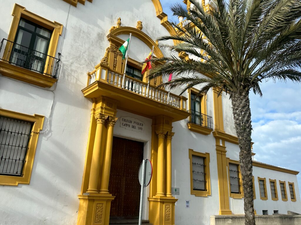 Colonial-style white building with ornate golden trim, arched doorway, balcony, and palm tree in Spanish plaza