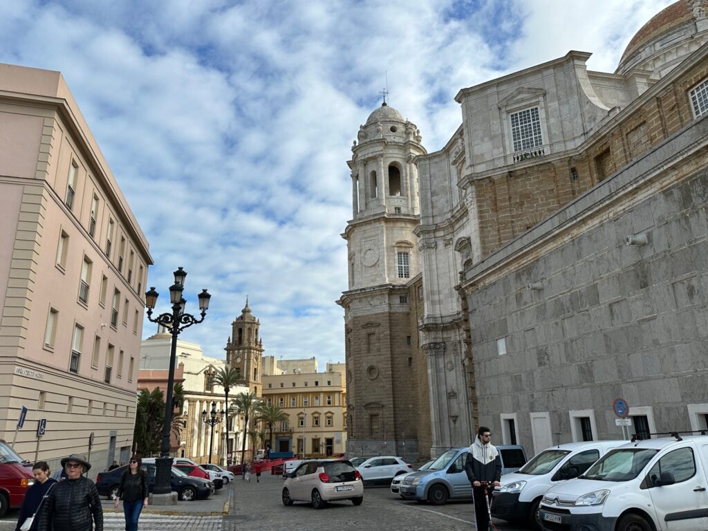 Historic European plaza featuring baroque cathedral with bell tower, surrounding classical buildings, parked cars, and pedestrians on sunny day