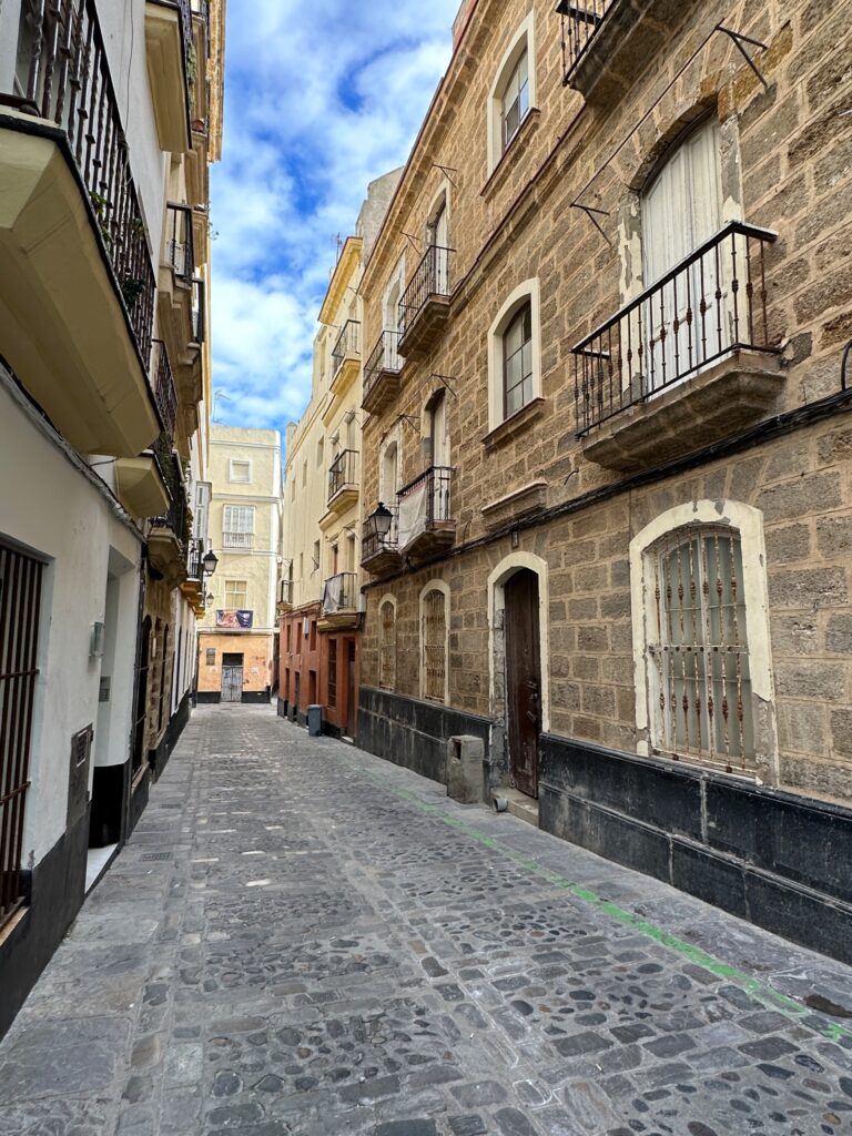 Narrow cobblestone alley lined with historic stone buildings, wrought iron balconies, and wooden doors under blue sky