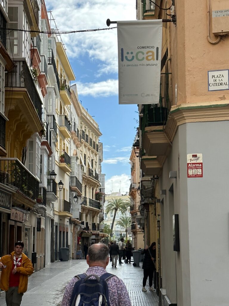 A pedestrian walks down a narrow European street lined with historic buildings, palm trees visible in the distance near a cathedral plaza
