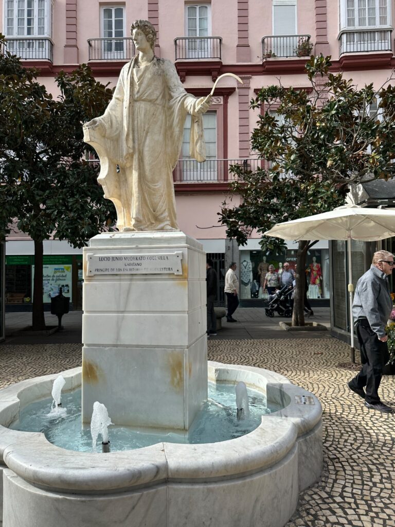 Classical white marble statue of a robed figure in a public plaza fountain, with pink buildings and pedestrians visible in the background