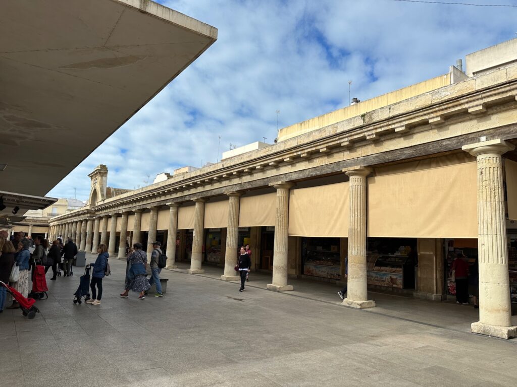 Classical colonnade with fluted columns beneath a modern concrete overhang, featuring people walking through a public plaza on a sunny day