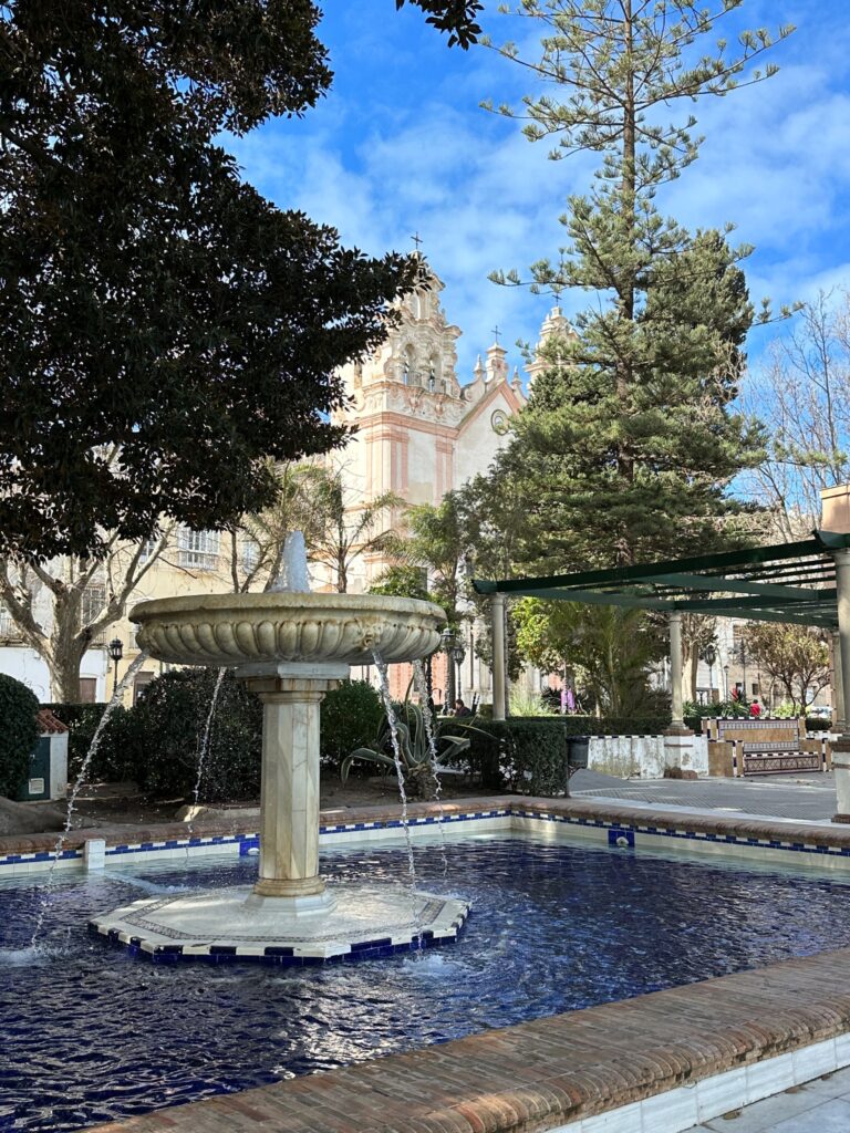 A decorative fountain in a Spanish plaza with a baroque church featuring pink domes and crosses in the background