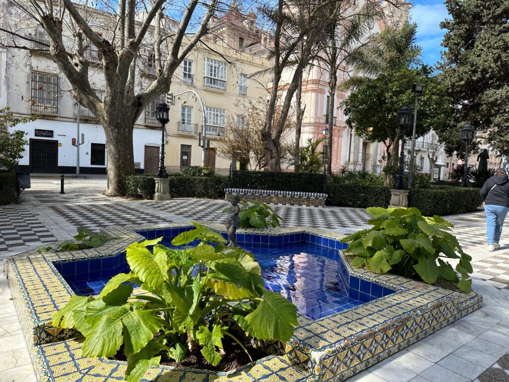 Blue tiled fountain with green plants in a Spanish plaza surrounded by historic buildings and trees
