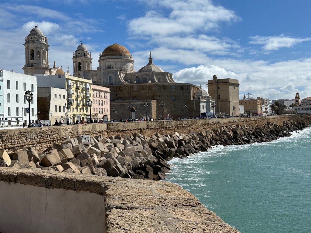 Coastal city with historic cathedral and bell towers overlooking turquoise Mediterranean waters and rocky breakwater