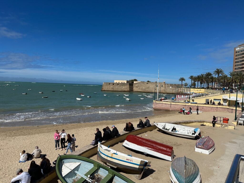 Sandy beach with moored fishing boats, beachgoers, fortress in background, and palm trees lining the Mediterranean coastline