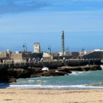Seaside harbor with historic stone pier, lighthouse, and beach with visitors on a sunny day