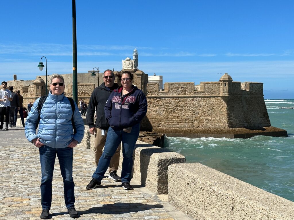 Three visitors stand on a stone pier beside a fortified coastal castle under blue skies