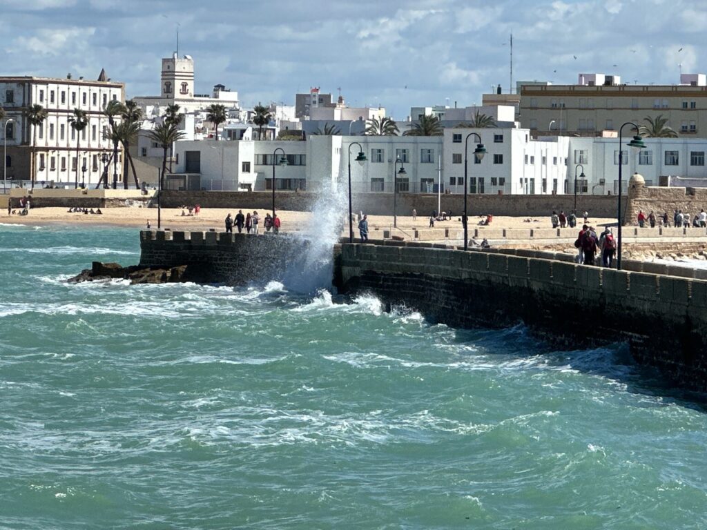 Waves crash against a stone pier with a sandy beach and white-washed buildings in a coastal town under blue sky