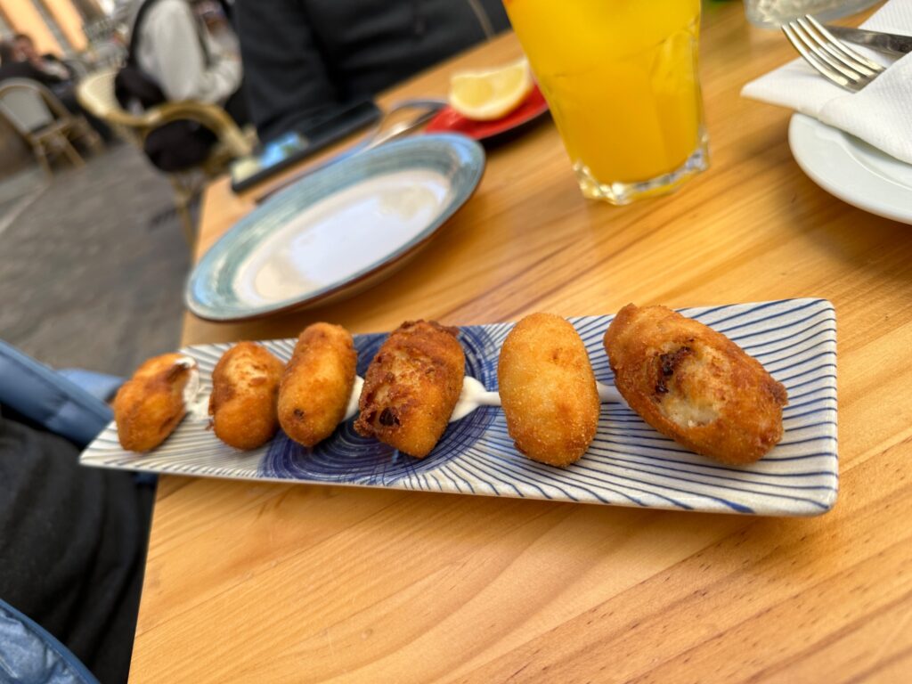 Six golden-brown croquettes arranged on a striped blue and white plate at a wooden dining table with place settings and orange juice