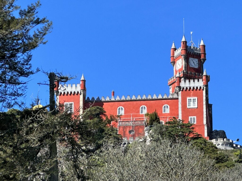 A striking red brick castle with towers and a clock perched on a rocky cliff against a clear blue sky