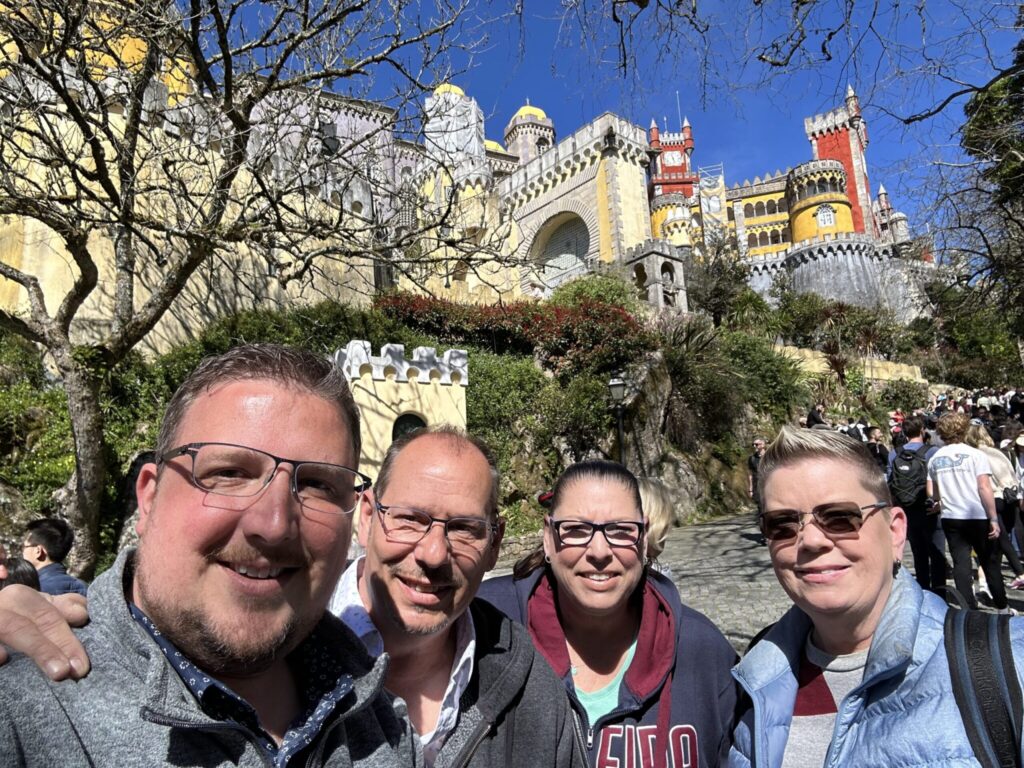 Four smiling people pose together in front of Pena Palace, a colorful castle with towers and walls on a hillside