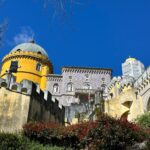 Colorful castle with yellow dome and pink tower surrounded by gardens under a bright blue sky