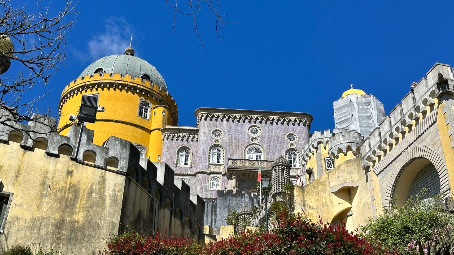 Colorful castle with yellow dome and pink tower surrounded by gardens under a bright blue sky