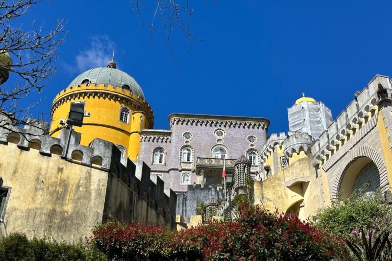 Colorful castle with yellow dome and pink tower surrounded by gardens under a bright blue sky