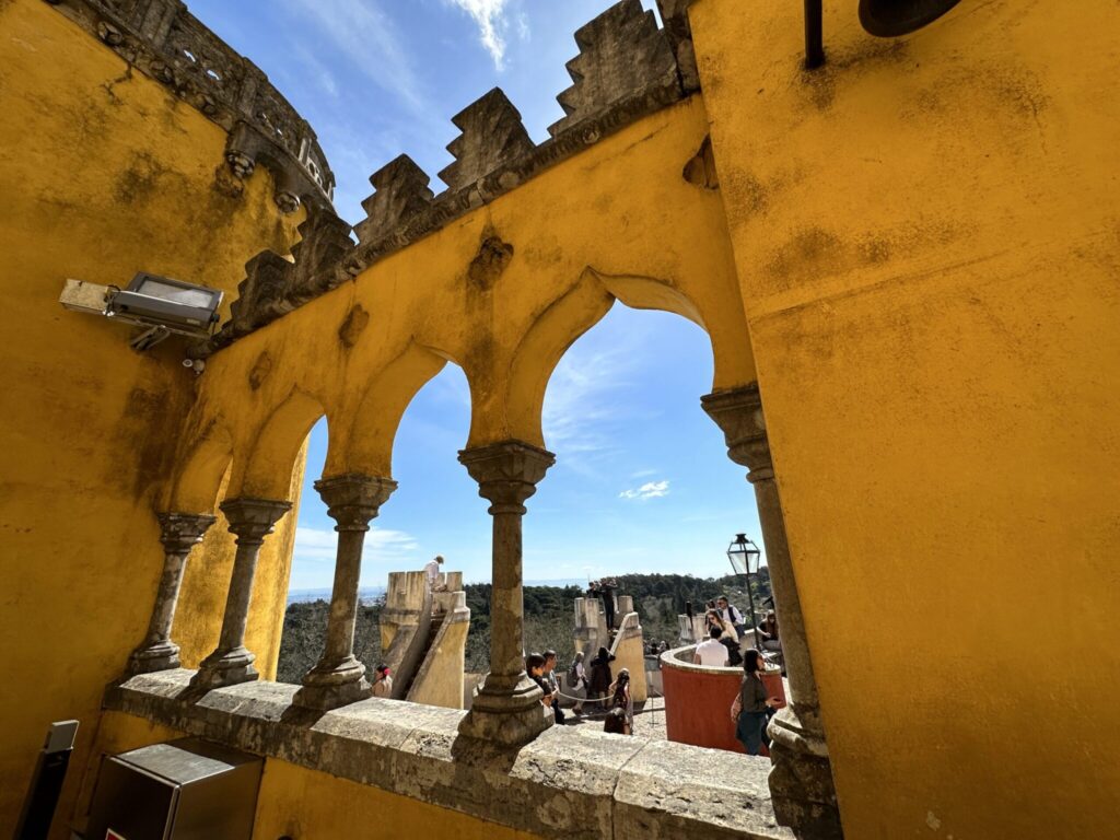 Yellow stone archway with ornate columns frames a hilltop castle and courtyard with visitors below