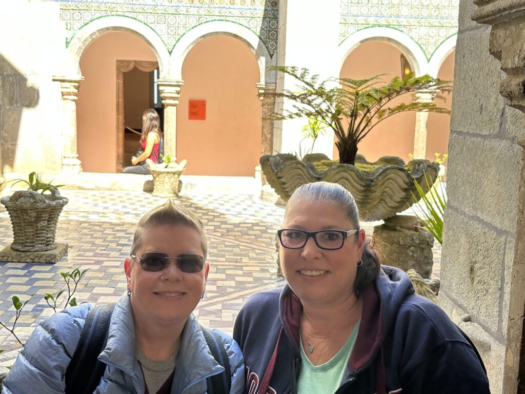 Two smiling visitors pose together in a sunlit Mediterranean courtyard with arched doorways and decorative tilework