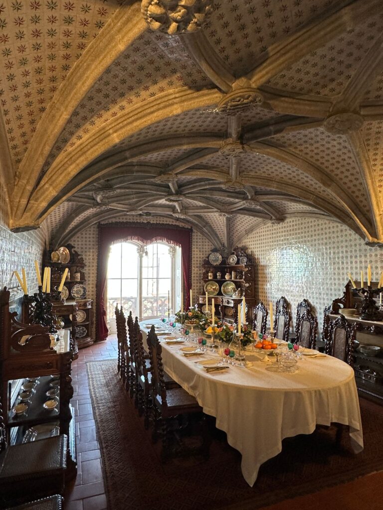 An elegant Gothic dining room features a long table with white tablecloth, ornate wooden chairs, decorative plates, and vaulted ceiling with star patterns
