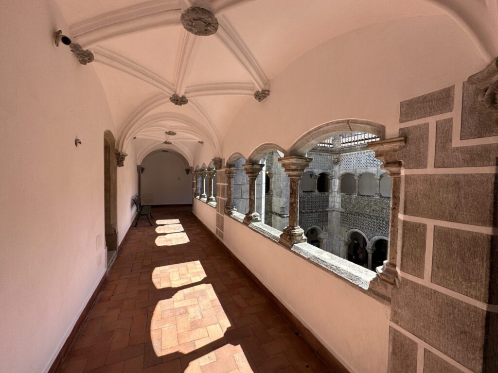 A long arched corridor with terracotta floor tiles, pink walls, and a cloister overlooking a courtyard with columns