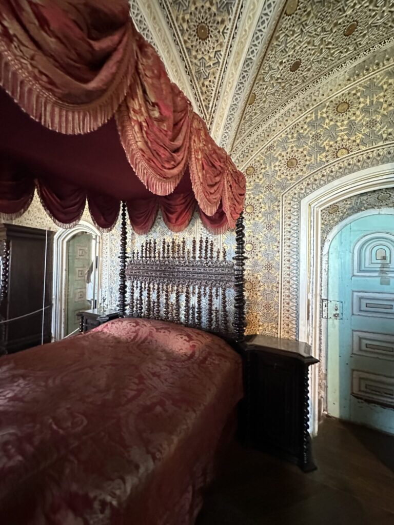 Ornate bedroom featuring a four-poster bed with burgundy canopy, decorative white and black beaded headboard, intricate patterned wallpaper, and turquoise doors