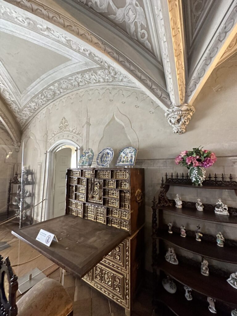 Ornate museum room displaying decorative furniture, porcelain plates, and collectibles beneath elaborate ceiling moldings and arches
