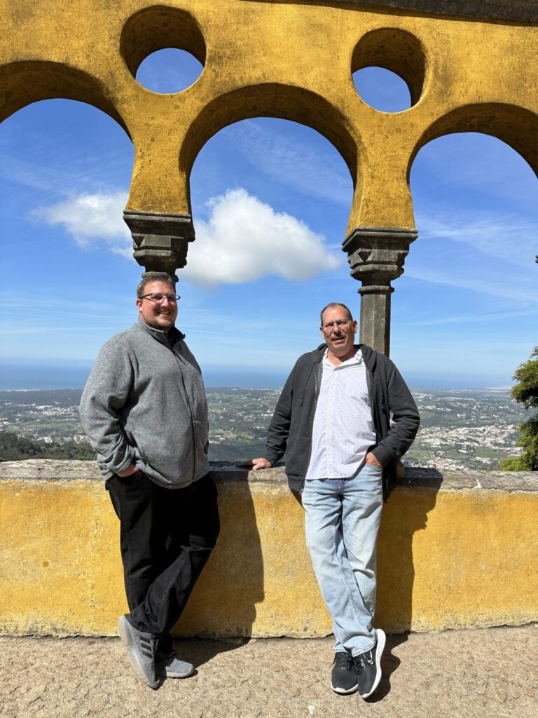 Two men stand beneath yellow stone arches with a coastal landscape and blue sky visible behind them
