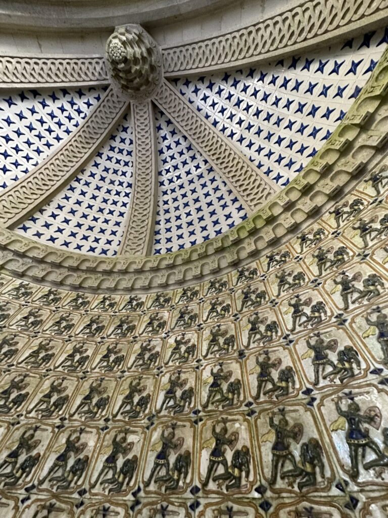 Ornate domed ceiling featuring blue stars, decorative rosette, classical figures in relief panels, and intricate geometric patterns