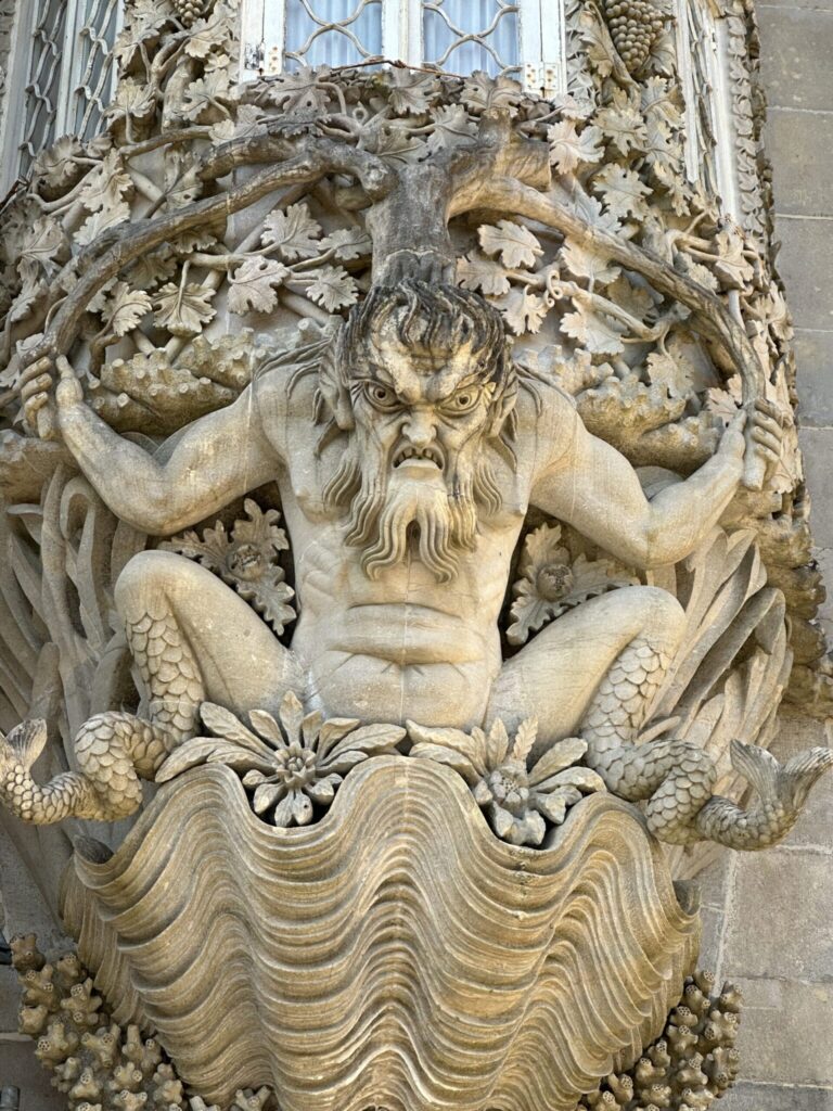 Stone carved ornamental head with beard and scaly creatures, featuring decorative foliage and wavy base elements on architectural facade