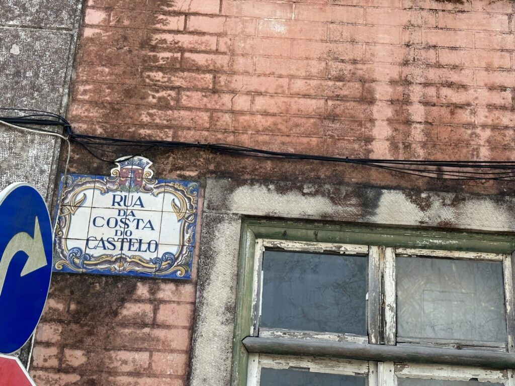 Portuguese street sign reading "Rua da Costa do Castelo" mounted on weathered brick wall beside green-framed window