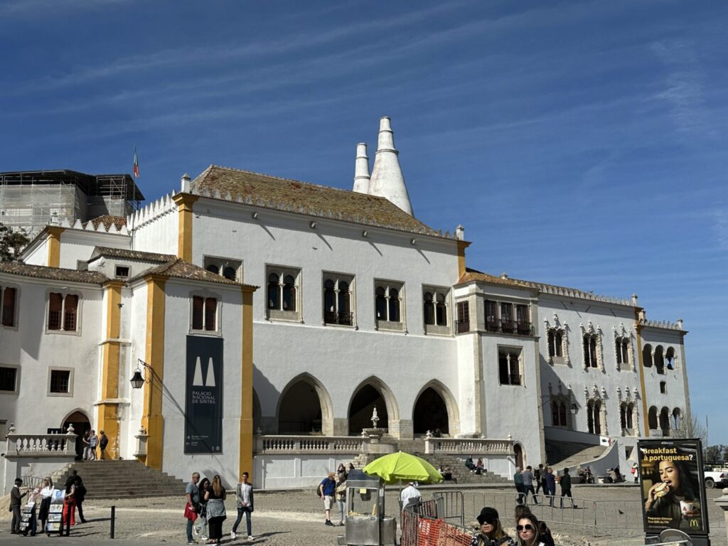 White stone palace with distinctive twin chimneys and Gothic arches surrounded by tourists under blue sky