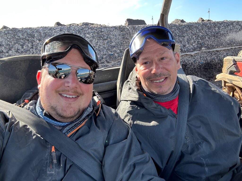 Two smiling men wearing winter jackets and ski goggles sit together in an open-air vehicle with a large recycling or waste facility visible behind them