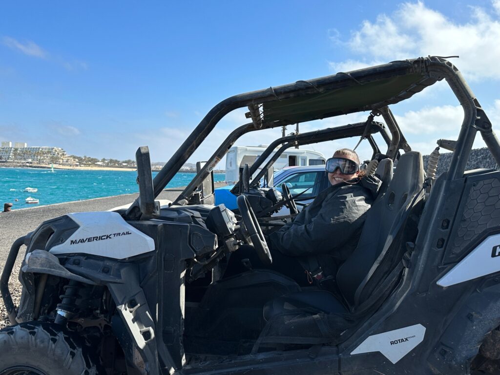 Person sitting in a white and black Maverick Trail buggy on a beach with turquoise water and coastal town visible behind