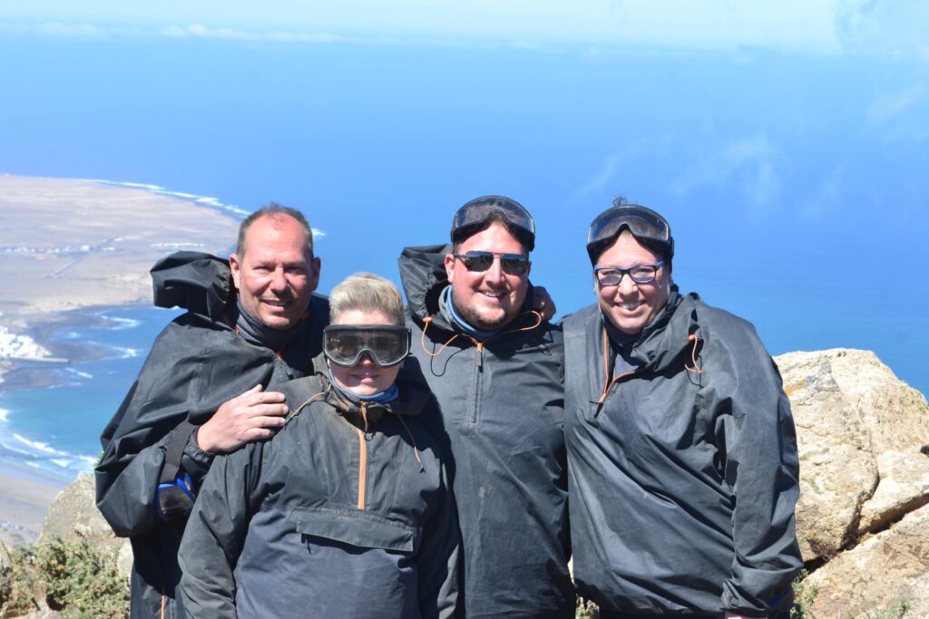 Four people in ponchos pose together on a mountain peak with a scenic valley and blue sky visible below