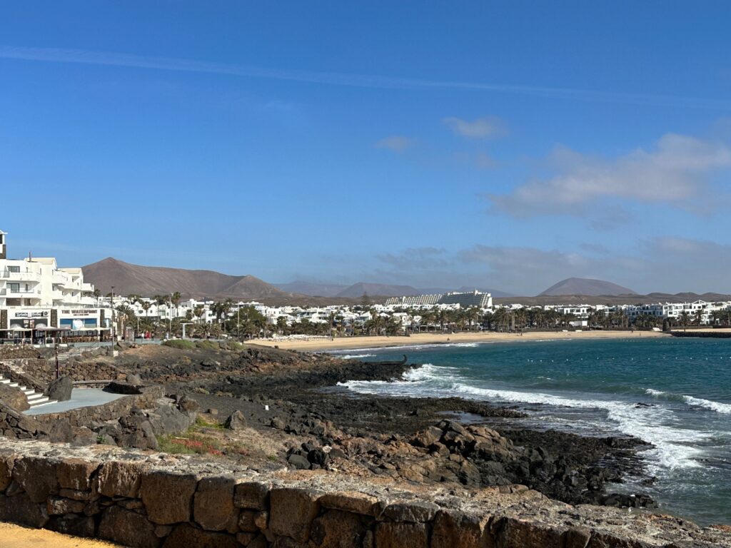 Coastal beach town with black volcanic rocks, turquoise waters, and arid mountains under blue sky