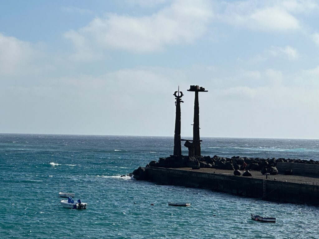 Two tall crosses stand silhouetted on a rocky breakwater extending into turquoise ocean waters under a partly cloudy sky
