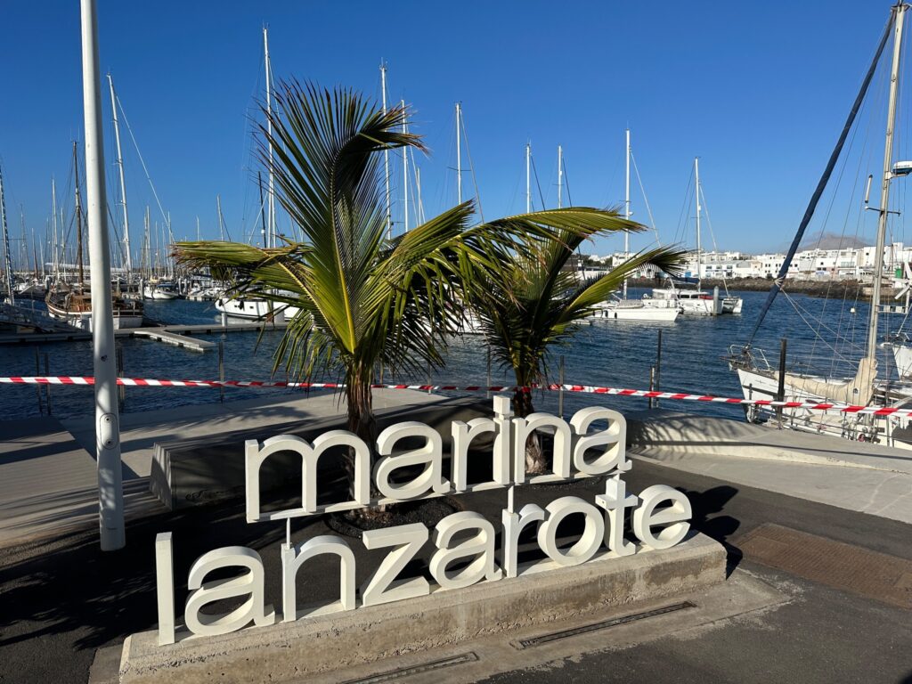 White "marina lanzarote" sign with palm tree beside sailboat-filled harbor under clear blue sky