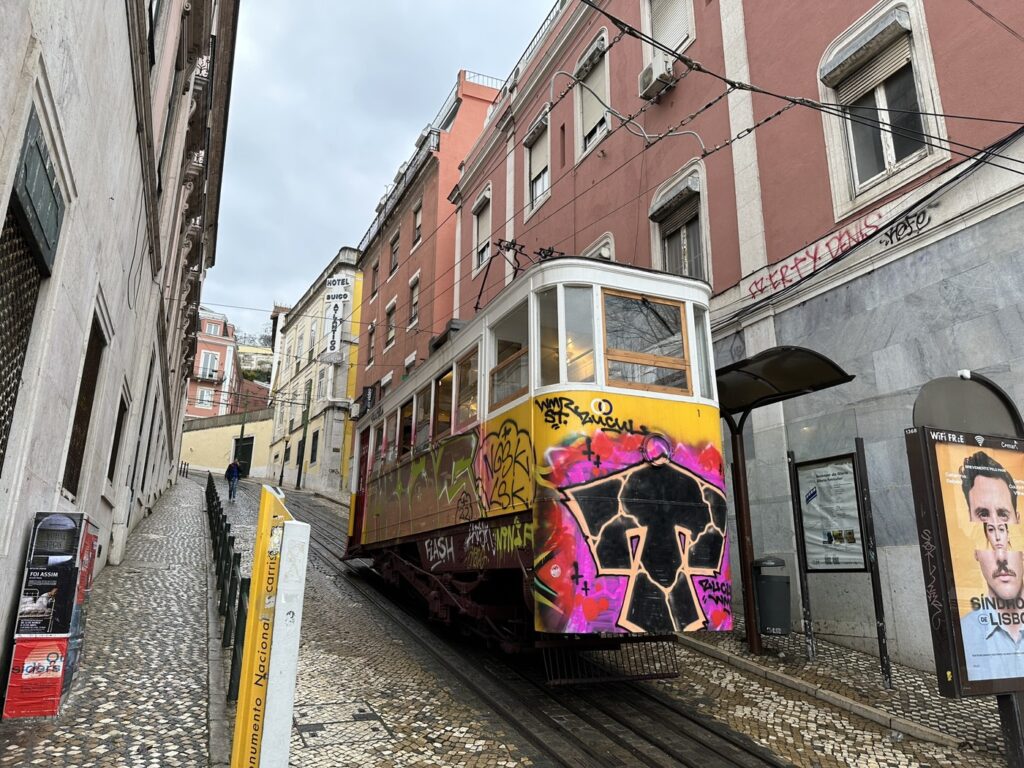 Yellow tram decorated with colorful graffiti art travels down a steep cobblestone street in historic Lisbon