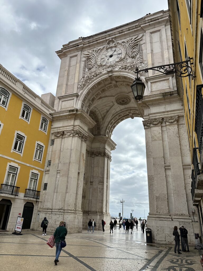 Ornate stone archway with clock tower in historic Lisbon plaza, featuring yellow buildings and people walking through