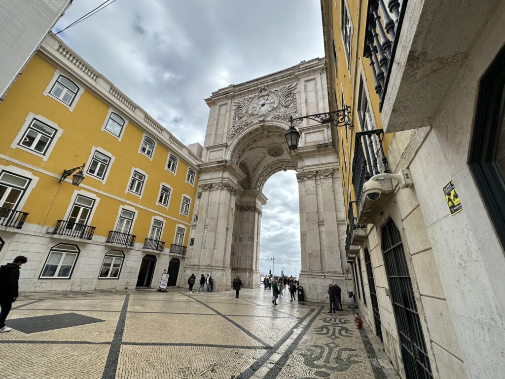 A grand stone archway frames a historic plaza in Lisbon, with a yellow building and traditional Portuguese pavement visible