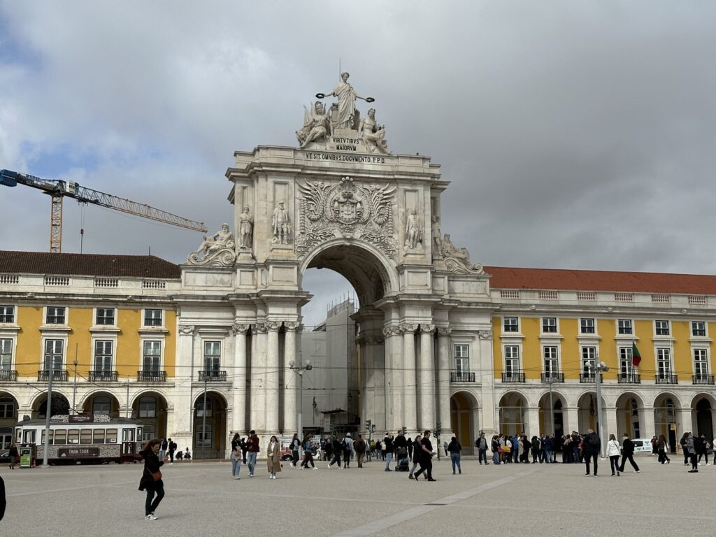 Historic triumphal arch with classical columns stands in a European plaza surrounded by yellow buildings and tourists