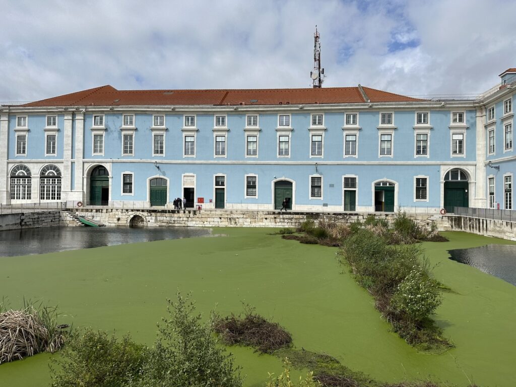Light blue colonial building with green doors beside a murky green waterway under cloudy skies