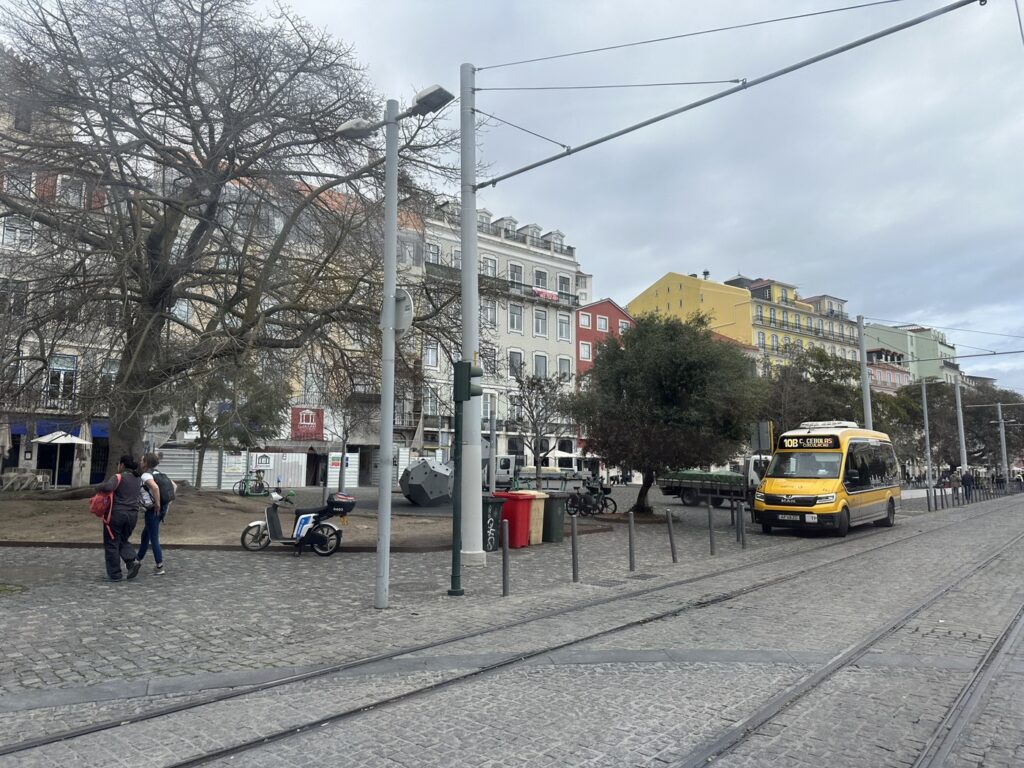 Yellow tram passes through a European city square with colorful buildings, bare trees, and pedestrians on a cobblestone plaza