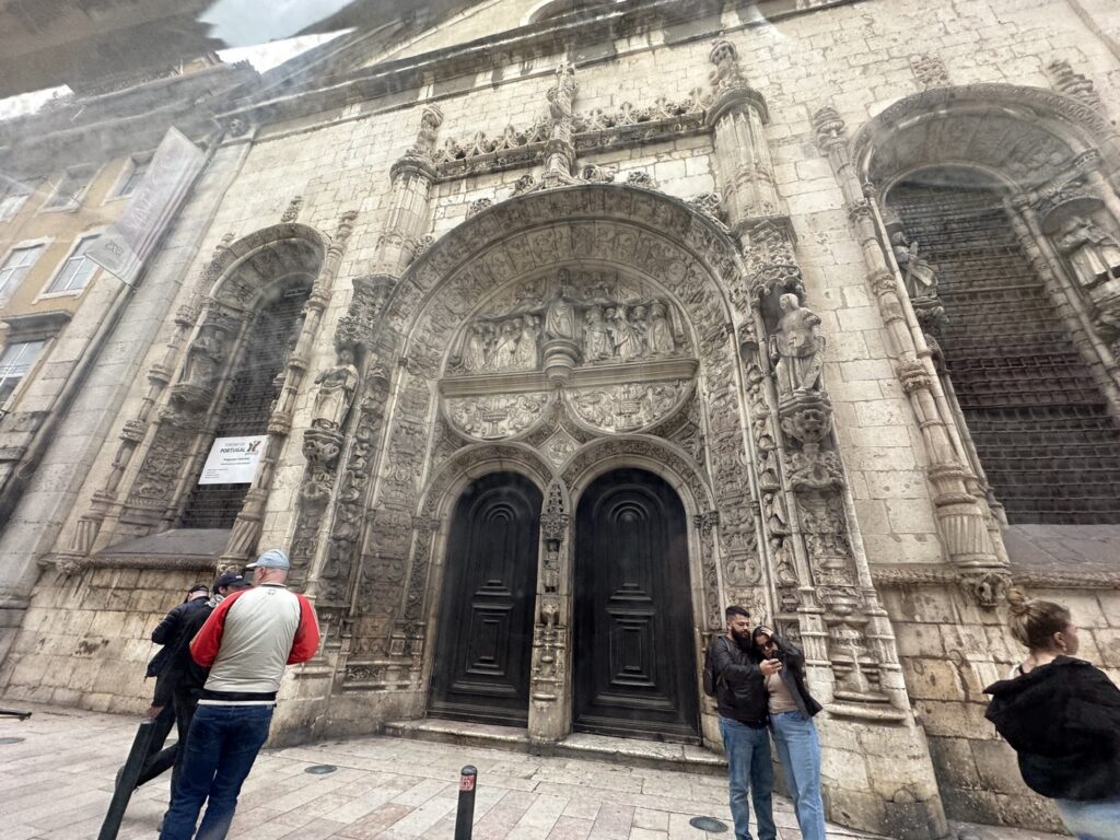 Ornate stone church entrance with elaborate carved Gothic archway, black double doors, and decorative sculptures, with visitors standing in front